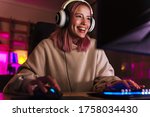 Image of joyful girl in headphones smiling and playing video game on computer while sitting at table indoors