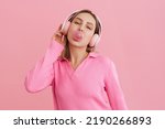 Small photo of Young cute girl in pink headphones blowing bubble gum over isolated pink background