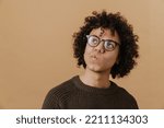 Small photo of Young handsome curly thoughtful boy in glasses with puckered lips looking aside, while standing over isolated beige background
