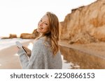 Beautiful young smiling woman wearing casual sweater using smartphone during walk at sunny beach