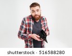 Upset young bearded man in plaid shirt holding empty wallet over white background