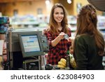 Image of smiling young lady standing in supermarket shop near cashier's desk holding credit card. Looking aside.