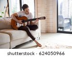 Pretty young man playing guitar while sitting on sofa in light living room