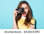 Close up portrait of a smiling pretty girl in dress taking photo on a retro camera isolated over blue background