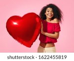 Happy girl holds red heart shape balloon. Photo of smiling young girl in love on pink background. Valentine's Day