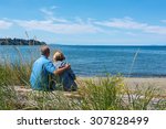 Adult man & woman sitting close to each other & hugging while looking out to the ocean, in Parksville, British Columbia, Canada