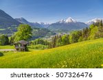 Panoramic view of idyllic mountain scenery in the Alps with traditional mountain chalet and fresh green mountain pastures with blooming flowers on a sunny day with blue sky and clouds in summer