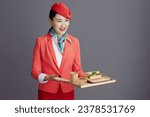 Small photo of smiling stylish air hostess asian woman in red skirt, jacket and hat uniform with a tray of food isolated on gray background.