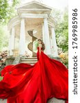 Small photo of Fashion Model Waving Long Fluttering Red Dress, Woman in Garden, Old White Alcove in Flowers, Outdoor Beauty Portrait