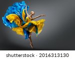 Beautiful Woman Ballet Dancer Jumping in Air in Colorful Fluttering Dress. Graceful Ballerina Dancing in Yellow Blue Gown over Gray Studio Background