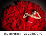 Small photo of Fashion Model in Red Fluffy Cloud Dress lying down. Beauty Woman Portrait with Silver Jewelry. Elegant Lady in Ball Gown looking a side over Gray Studio Background