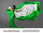 Woman dancing with Nigerian Flag in Green White Dress. Fashion Model with Hair in Long Gown with flying Silk Scarf. Waving Chiffon Fabric over Gray. Nigeria Independence Day