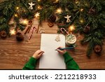Letter to Santa. Christmas Child Hands Close up writing Xmas Gifts Shopping Wish List over brown Wooden Table. Wood Background with Fir Tree Branch decorated Christmas Lights and Toys