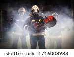Small photo of Group of professional firefighters wearing full equipment, oxygen masks, and emergency rescue tools, circular hydraulic and gas saw, axe, and sledge hammer. smoke and fire trucks in the background.