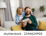 Small photo of Happy Woman Presenting Gift To Husband. Excited Young Woman Closed Eyes Presenting Good Unexpected Present To Husband At Home. Loving Girlfriend Making Romantic Surprise To Boyfriend