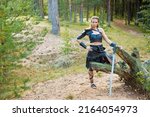 Small photo of Pretty brigand girl wearing black leather shoulder guard, skirt, lorica and bracer with steel estramacon is resting on the fallen tree in a summer forest
