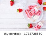 Plate of homemade strawberry vanilla yogurt popsicles. Above view corner border on a white wood background with copy space.