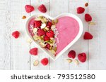 Healthy raspberry smoothie in a heart shaped bowl with superfoods. Above scene on a white wood background.