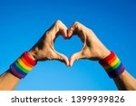 Small photo of Gay athlete making hand heart with gay pride rainbow colors sport wristbands against bright blue sky