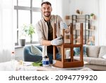 Small photo of furniture renovation, diy and home improvement concept - happy smiling man sanding old wooden table or chair with sponge
