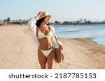 people, summer holidays and leisure concept - happy young woman in bikini swimsuit, white shirt and straw hat with bag walking along beach