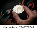 Hand holding a silver Ethereum (ETH) cryptocurrency coin with candle stick graph chart and digital background.