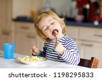 Small photo of Adorable baby girl eating from spoon noodle, pasta macaroni. Cute healthy toddler child, daughter with spoon sitting in highchair and learning to eat by itself in domestic kitchen or nursery