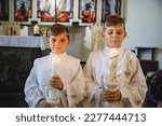 Small photo of Two little kids boys receiving his first holy communion. Happy children holding Christening candle. Tradition in catholic curch. Kids in a church near altar. Siblings, brothers in white gowns.