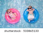 Two month old twin baby sister and brother sleeping on tiny, inflatable, pink and blue swim rings. They are wearing crocheted swimsuits and sunglasses.