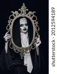 Small photo of Halloween. Portrait of a scary devilish nun with a picture frame in her hands on a black background. Horrors.