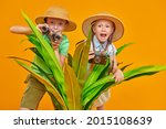 Young travelers, a girl and a boy, explore nature, they look out from behind palm leaves with binoculars and a camera. Summer vacation, tourism. Studio portrait on a yellow background. 