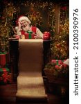 Small photo of Cheerful smiling Santa Claus thinks about a list of good and naughty children sitting in festively decorated room with a gift box and a long magic scroll in front of him. Christmas fairy tale.