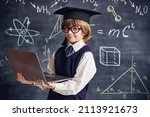 Portrait of a clever little boy in school uniform and glasses holding a laptop in the background of a blackboard with scientific formulas and diagrams. Smart children. Education. 