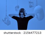 Small photo of Mental disorders. A girl in black clothes holds in despair her face with her hands surrounded by different masks in a room with blue lighting. Human roles. Hypocrisy.