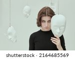 Small photo of Theater of life. A girl in black clothes stands in a white room surrounded by various masks looking intently at the camera. Hypocrisy. Human roles. Mental disorders.