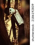 Small photo of Halloween. Portrait of a mysterious gentleman in a white carnival mask and a bowler hat peeking out from behind a tree in an old autumn park. Mime, carnival. Halloween. Vintage style.