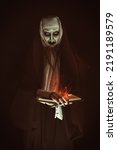 Small photo of An evil cursed nun with a burning bible and a cross in her hands looks at the camera on a black background. Halloween. Horrors.