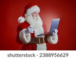 Modern Santa Claus looking at the laptop and is surprised. Red studio background. Christmas and New Year. Modern technologies.
