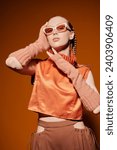 Small photo of Fashion model girl posing in stylish diamond-shaped glasses and an orange top on an orange studio background. Fashion and style for sunglasses. Summer haute couture collection.