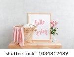 Frame with text HELLO SPRING, woven straw bag and vase with pink rose flowers on a wooden table on a background of light gray walls. Home interior decor.