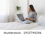 Young beautiful pregnant woman sitting on bed with laptop.