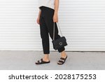 Woman wearing beige t-shirt, black pants, bag and flat sandals walking outdoor near white roller door. Details of stylish trendy basic minimalistic casual outfit. Street fashion. Women's legs, no face