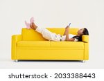 Smiling asian woman types text message on cell phone, enjoys online communication lying on yellow couch on white background. Technology, communication concept