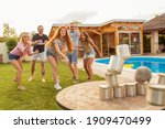 Small photo of Group of cheerful young friends having fun playing the knock down tin cans by throwing a ball game while at poolside summertime outdoor party