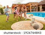 Small photo of Group of cheerful young friends having fun playing the knock down tin cans by throwing a ball game while at poolside summertime outdoor party