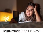 Cheerful young woman lying in bed at night, having video call using smart phone