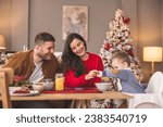 Young parents having breakfast with their toddler on Christmas morning at home, baby sitting in high chair holding spoon and stirring food
