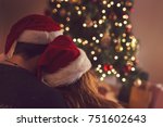 Couple in love sitting next to a Christmas tree, wearing Santa's hats, hugging and looking away from the camera towards the tree. Selective focus on the girl's hat