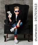 Small photo of Daring and sassy young rich boy kid millionaire sits in big luxury armchair and demonstrates bundle of money dollar bills