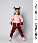 Small photo of Frolic smiling kid girl with straight brunette hair with buns in two-colored pink brown sport suit hoodie and pants poses with legs wide apart and hands on hips